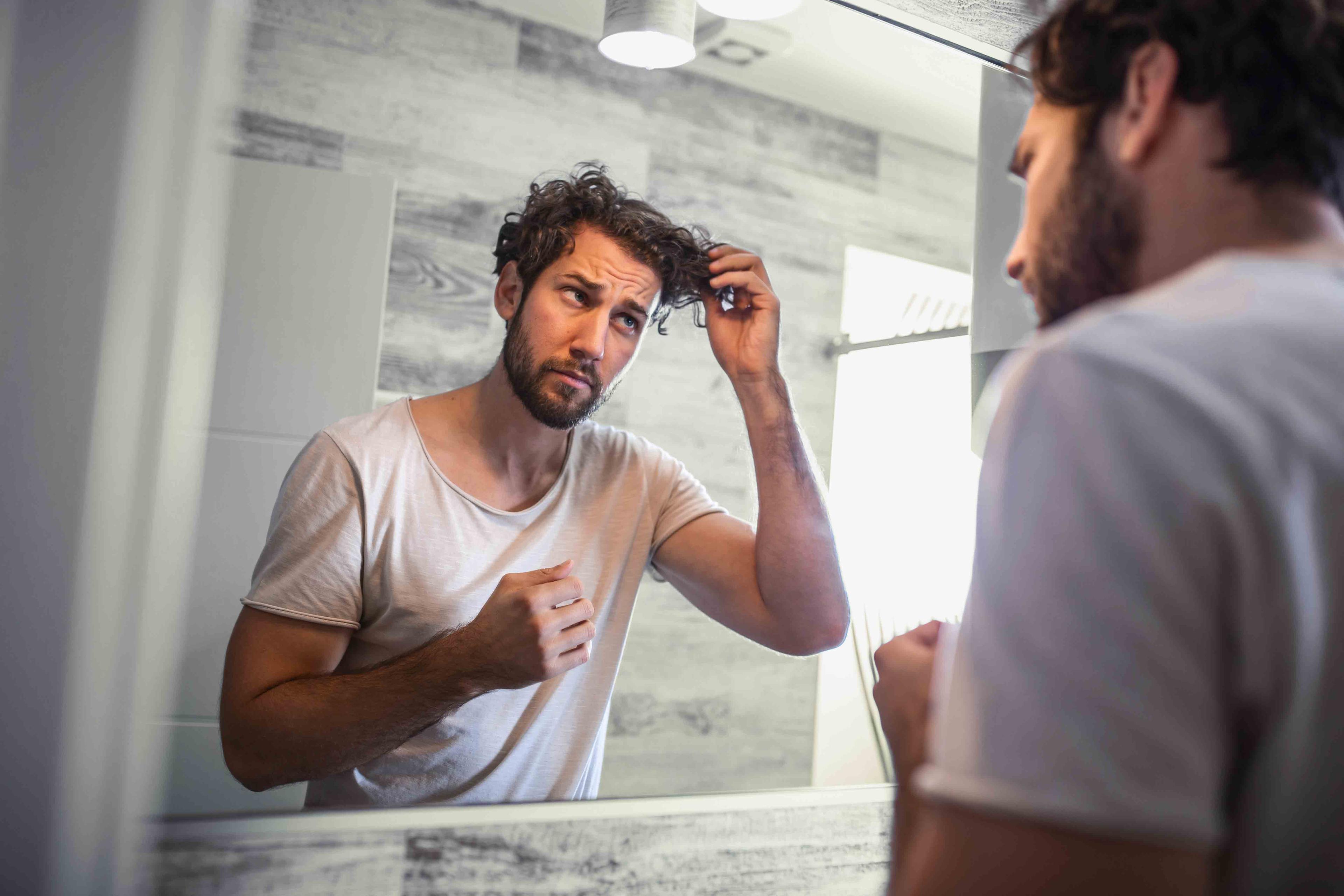 Young brunette man with beard touching hair while looking in bathroom mirror with concerned expression on his face.