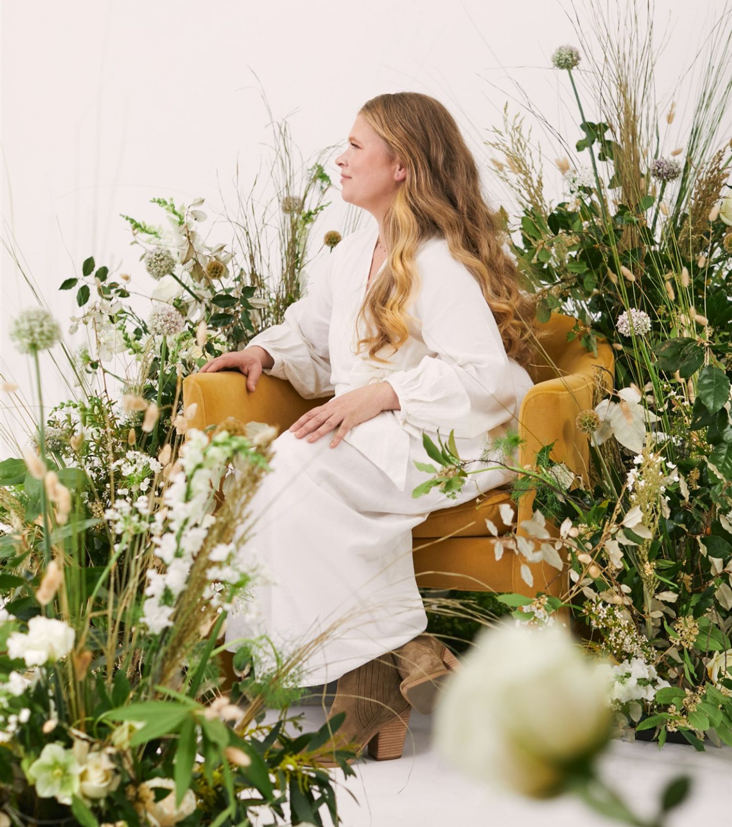 Woman with long wavy light brown hair sitting in profile in a gold armchair surrounded by lush tall wildflower arrangements