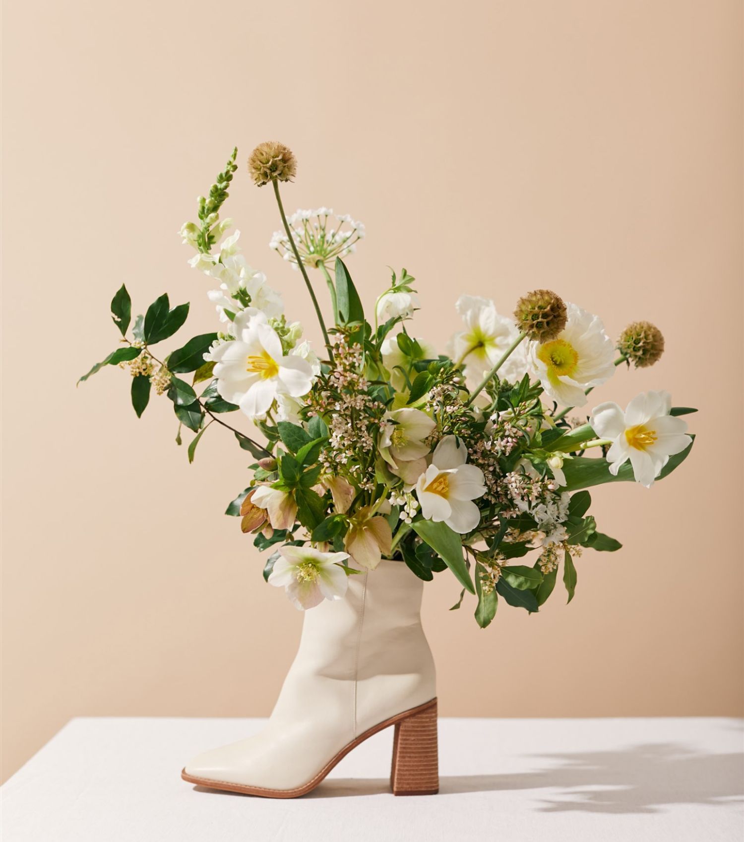 Bouquet of white and yellow wildflowers inside a short white boot with a stacked heel on a table with a beige background