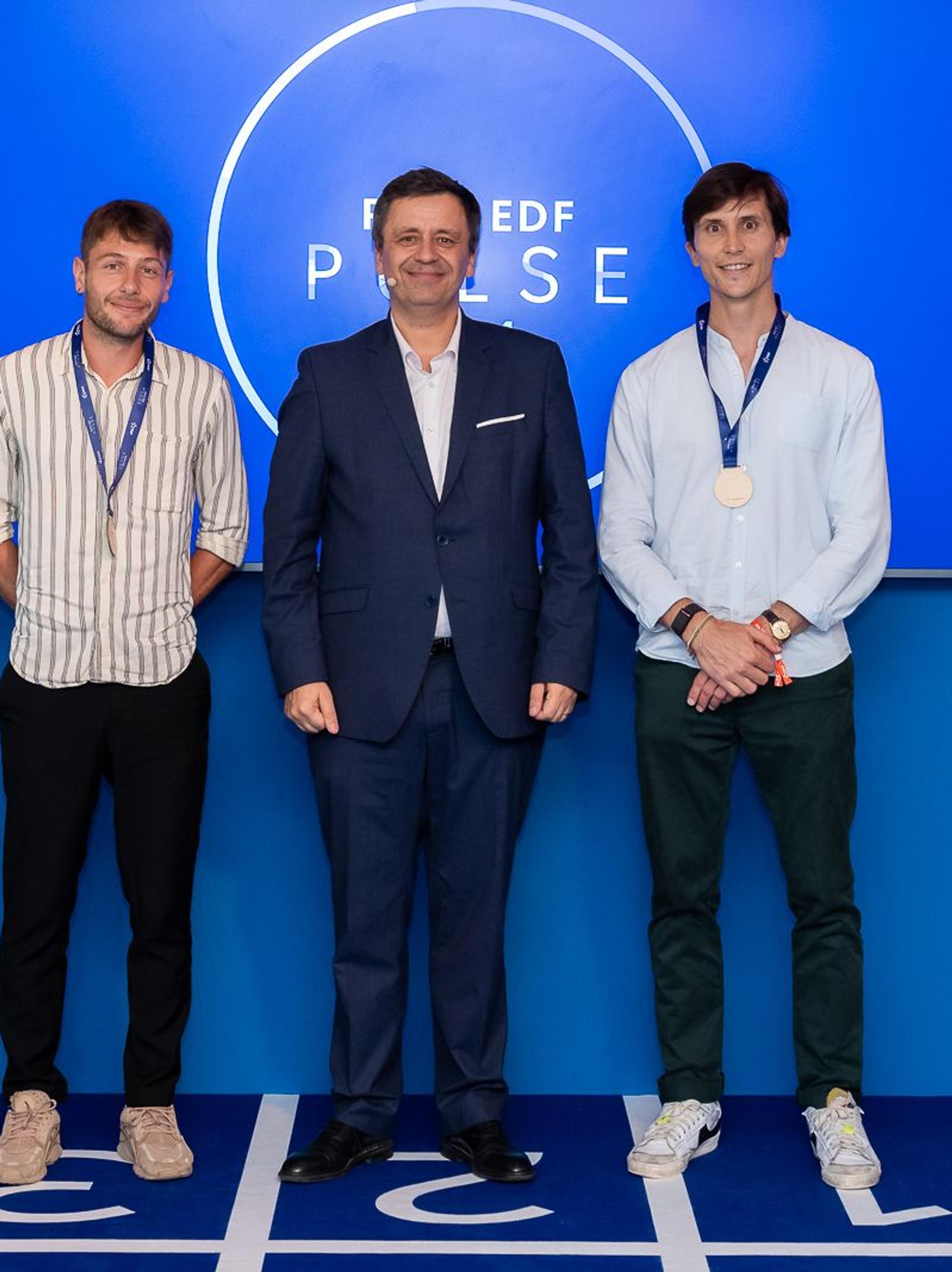 Aerleum team member stands on the winner's podium with a gold medal at the EDF Pulse Awards ceremony, alongside other finalists and an EDF representative, in front of a blue backdrop with the EDF Pulse logo.