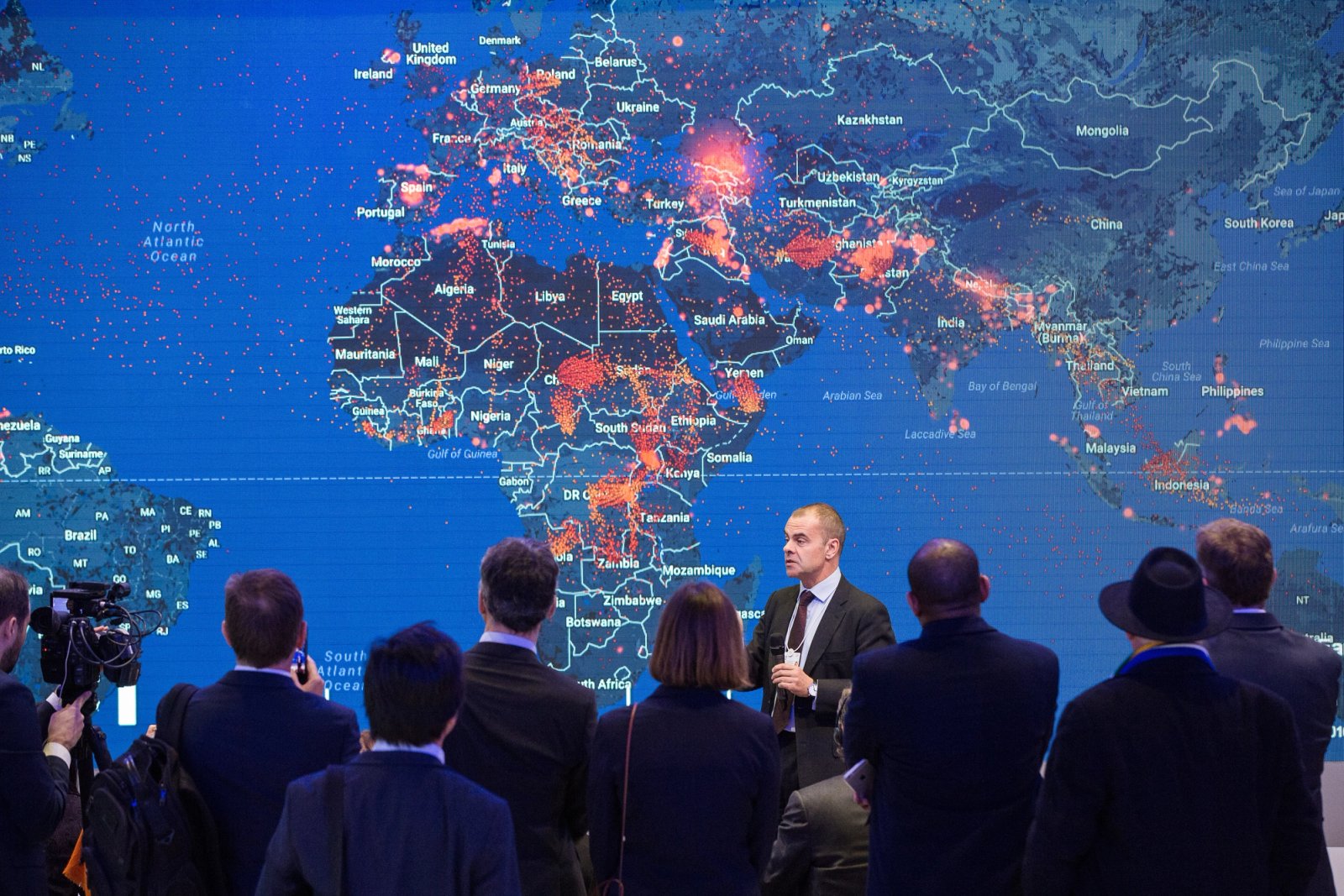 Man in a suit presenting to a group of attendees in front of a large digital world map display covered with red data points, suggesting global activity or cybersecurity monitoring.