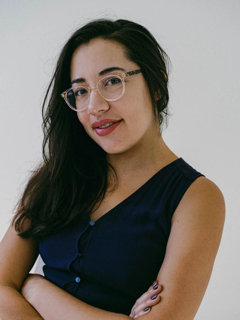 Image of Sarah Riazati in front of a white background wearing a navy blue sleeveless top with glasses and hair pulled to the side
