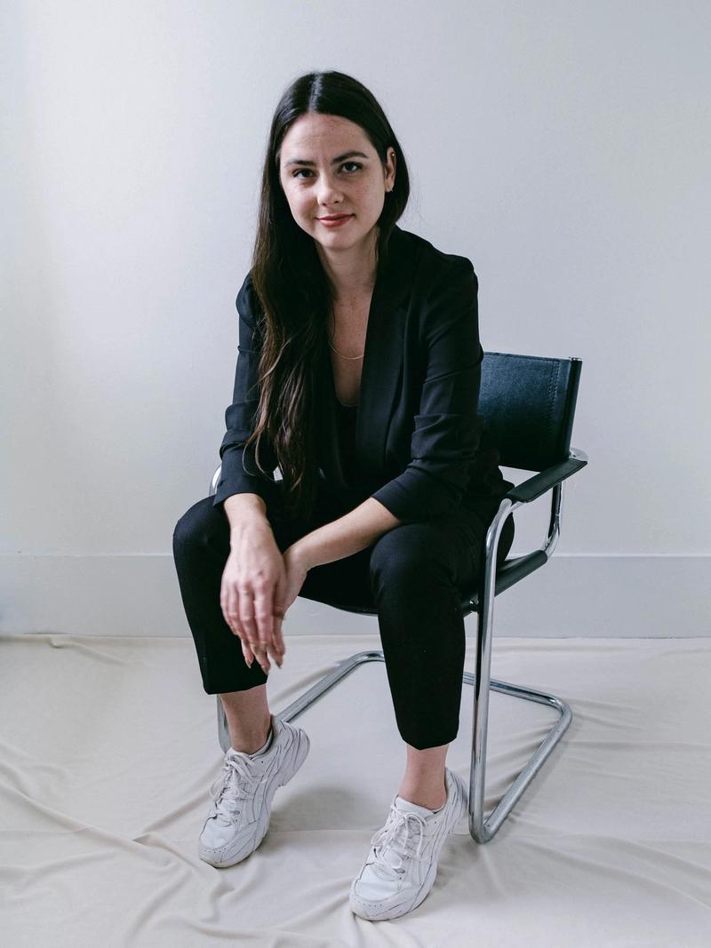Image of Hannah Lee in a black suit and white sneakers, leaning forward in a black and chrome chair.
