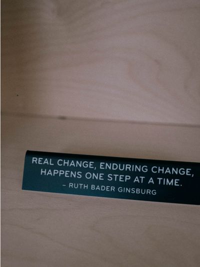 A black engraved plaque sits in a wooden bookshelf reading "Real change, enduring change, happens one step at a time" -Ruth Bader Ginsburg