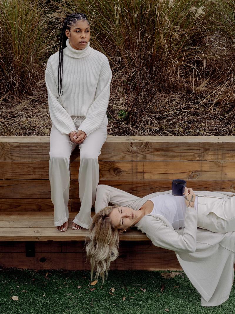 An image of two women on a wood bench. The woman on the left is in all white sitting on the top of the bench. The woman on the right is laying on the bench in all white with a coffee cup on her stomach.