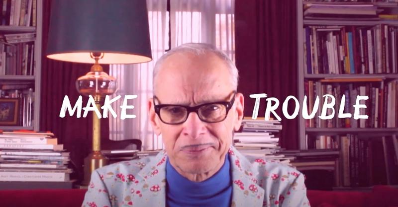 Image of a man with glasses, a blue turtleneck, and mushroom print blazer sitting in front of a widow with lamp and bookshelves in the background. White text laid over top saying Make Trouble