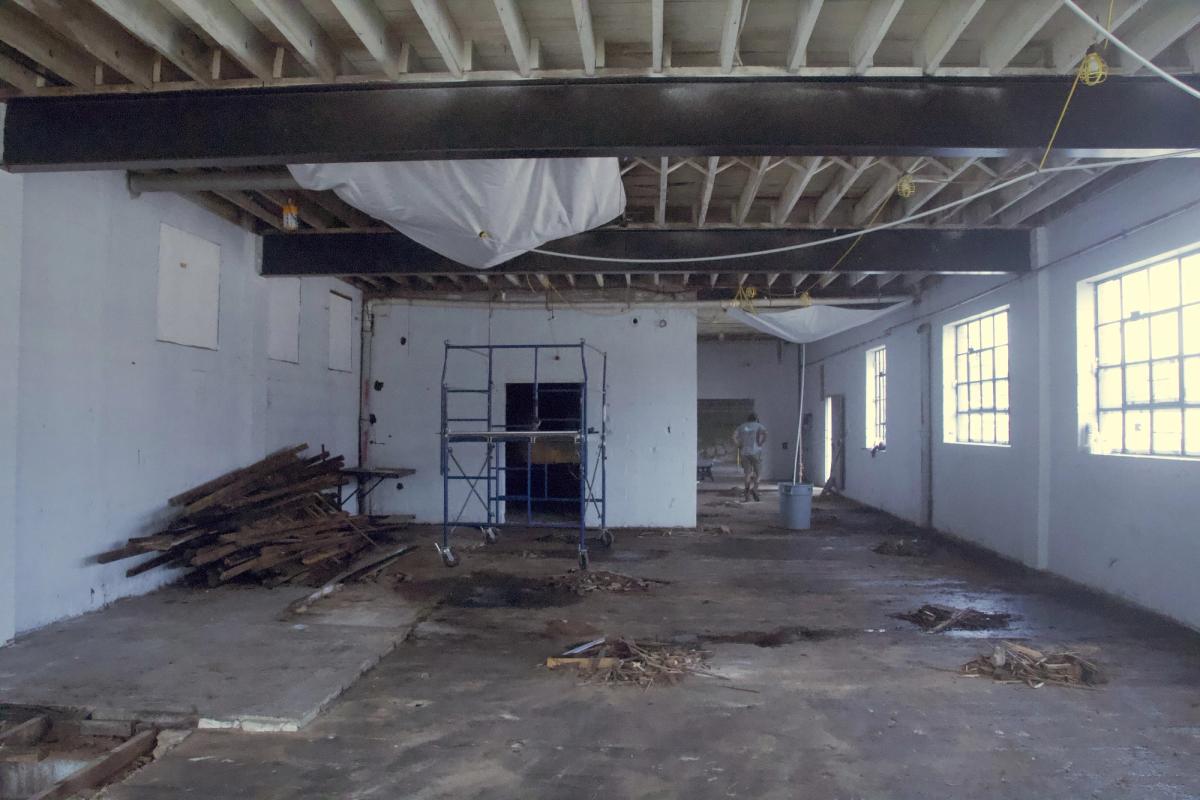Wide interior view of The Umbrella with exposed ceiling framing, a scaffold, and removed lumber stacked along a wall.