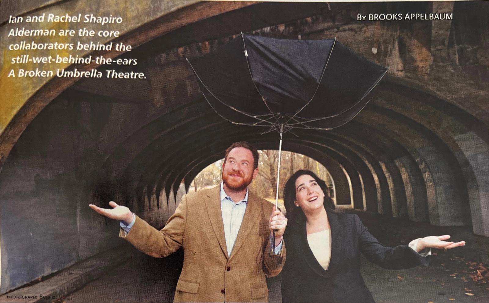 Ian Alderman and Rachel Shapiro Alderman stand beneath an inside-out umbrella in a stone tunnel, holding their free hands out to the sides.