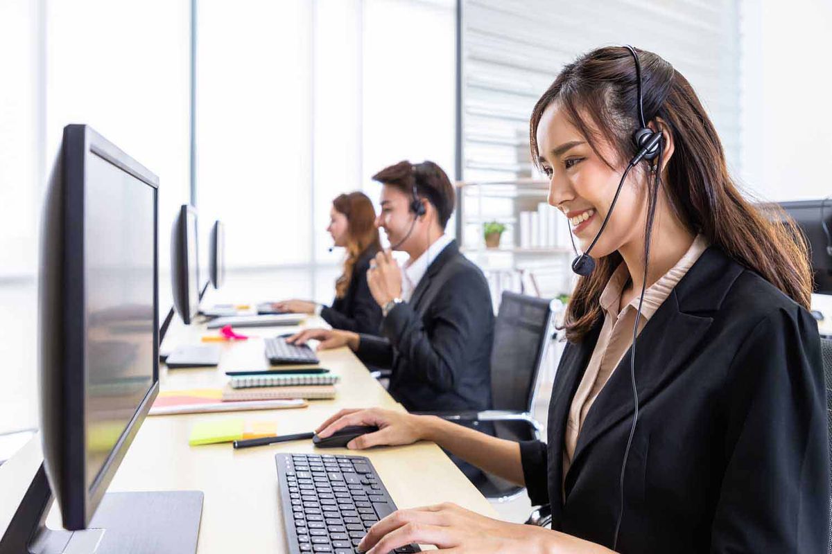 A smiling female retail customer service agent with headphones typing on a computer with her colleagues in an office.