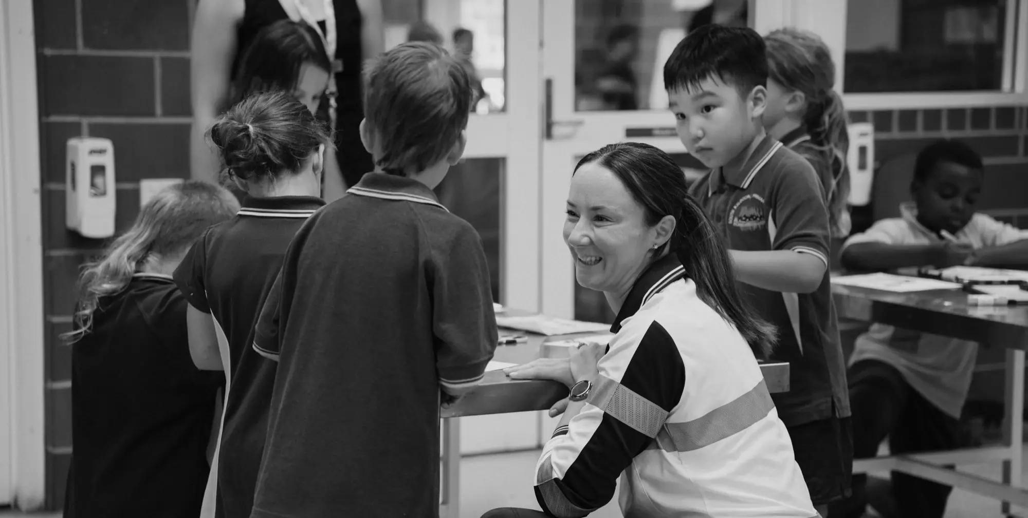 A teacher crouching beside primary school students working at a classroom bench.