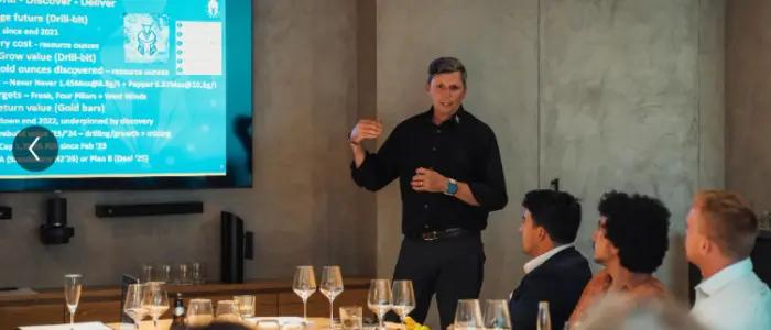 A speaker presenting results slides to guests seated at a long table set with wine glasses.