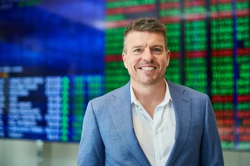 A man in a suit in front of a share price board showing green and red figures.