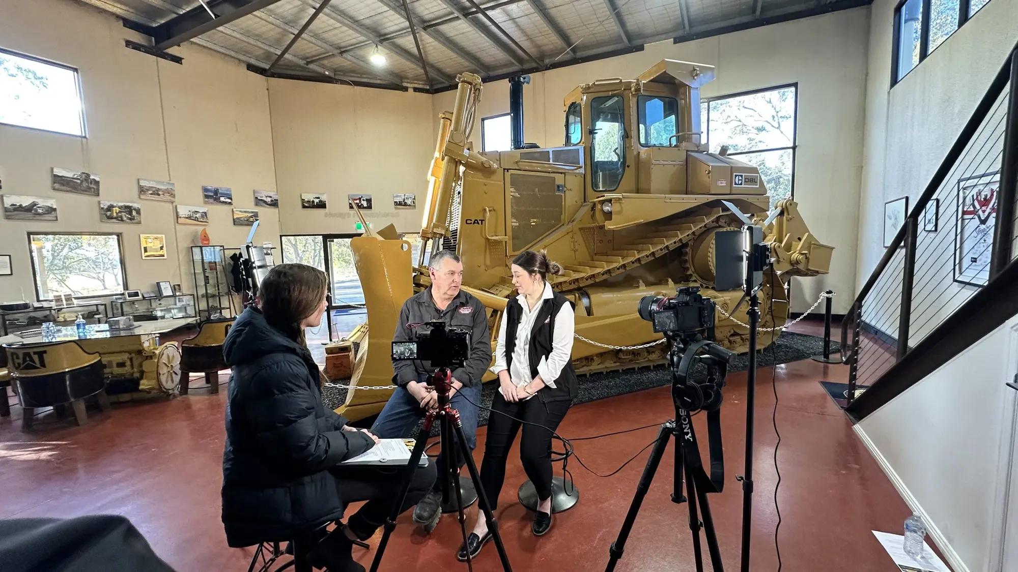 A film crew interviewing someone in front of a Caterpillar machine.