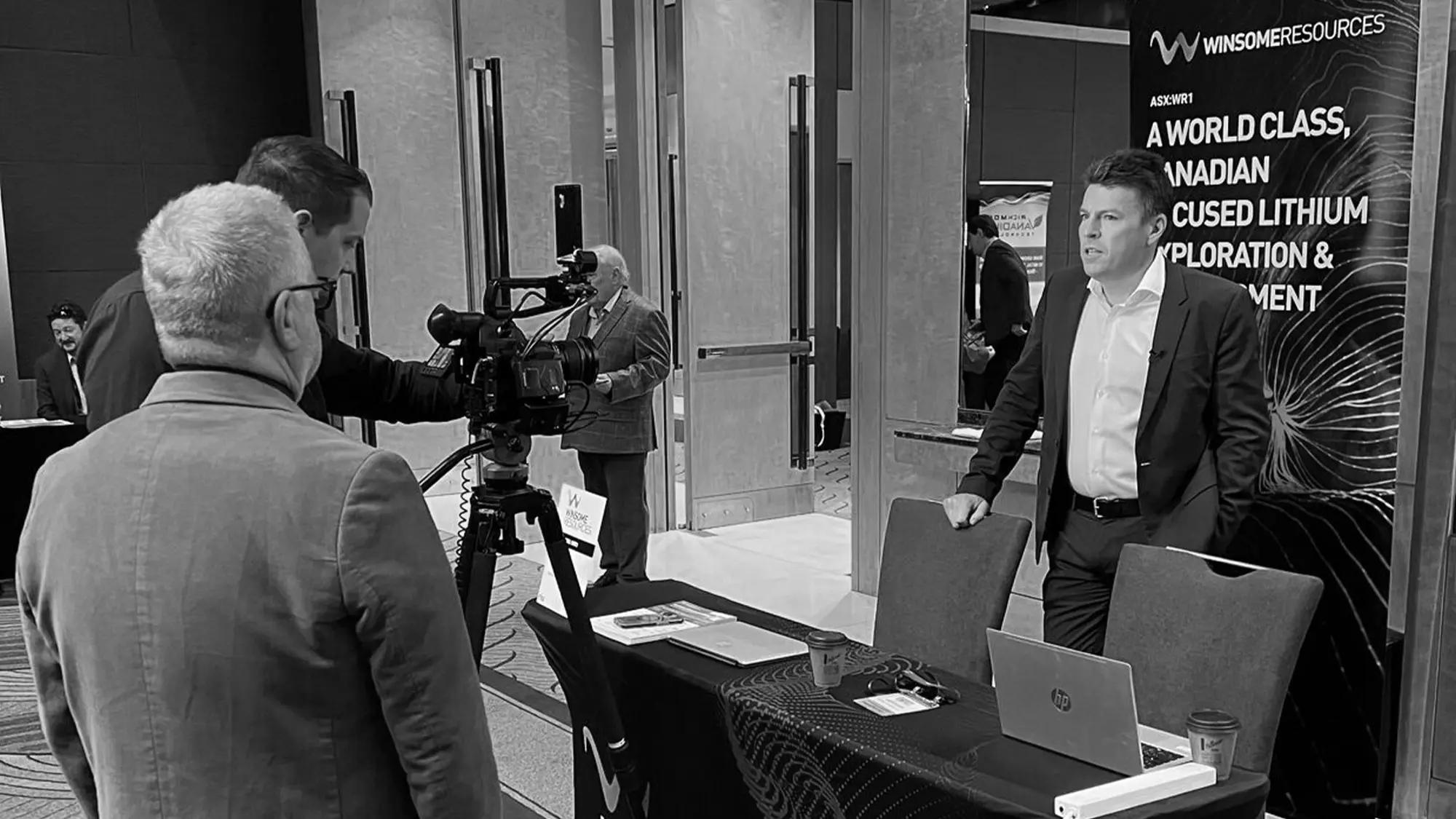 A camera operator filming a man beside a Winsome Resources banner at an investor event.