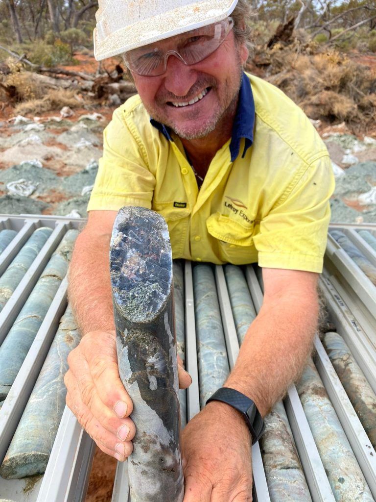 A geologist in a yellow shirt holding a length of drill core over a core tray.
