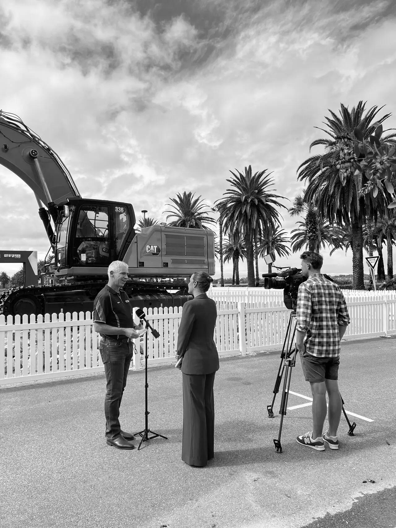A television crew interviewing a man in front of an excavator at an outdoor site.