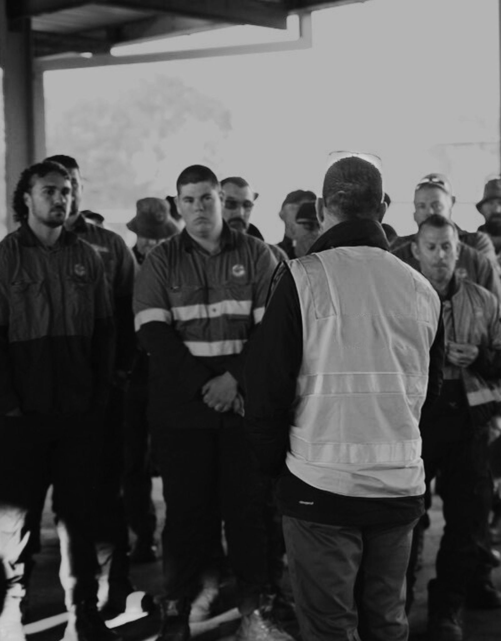 A worker in a hi-vis vest briefing a group of outdoor staff in uniform at a depot.