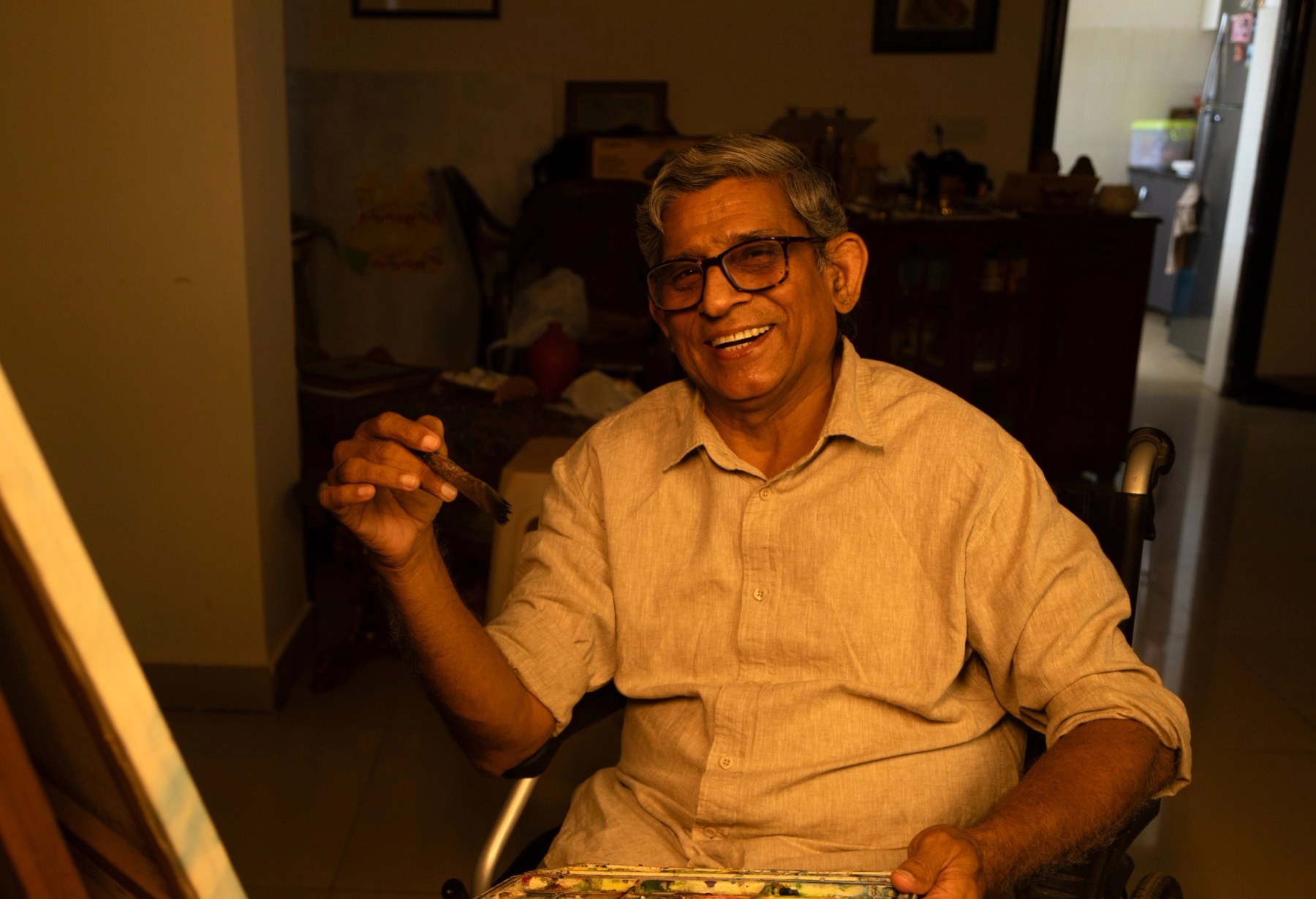 A smiling older man in a wheelchair painting at an easel, brush in hand and a well-used watercolor palette in his lap
