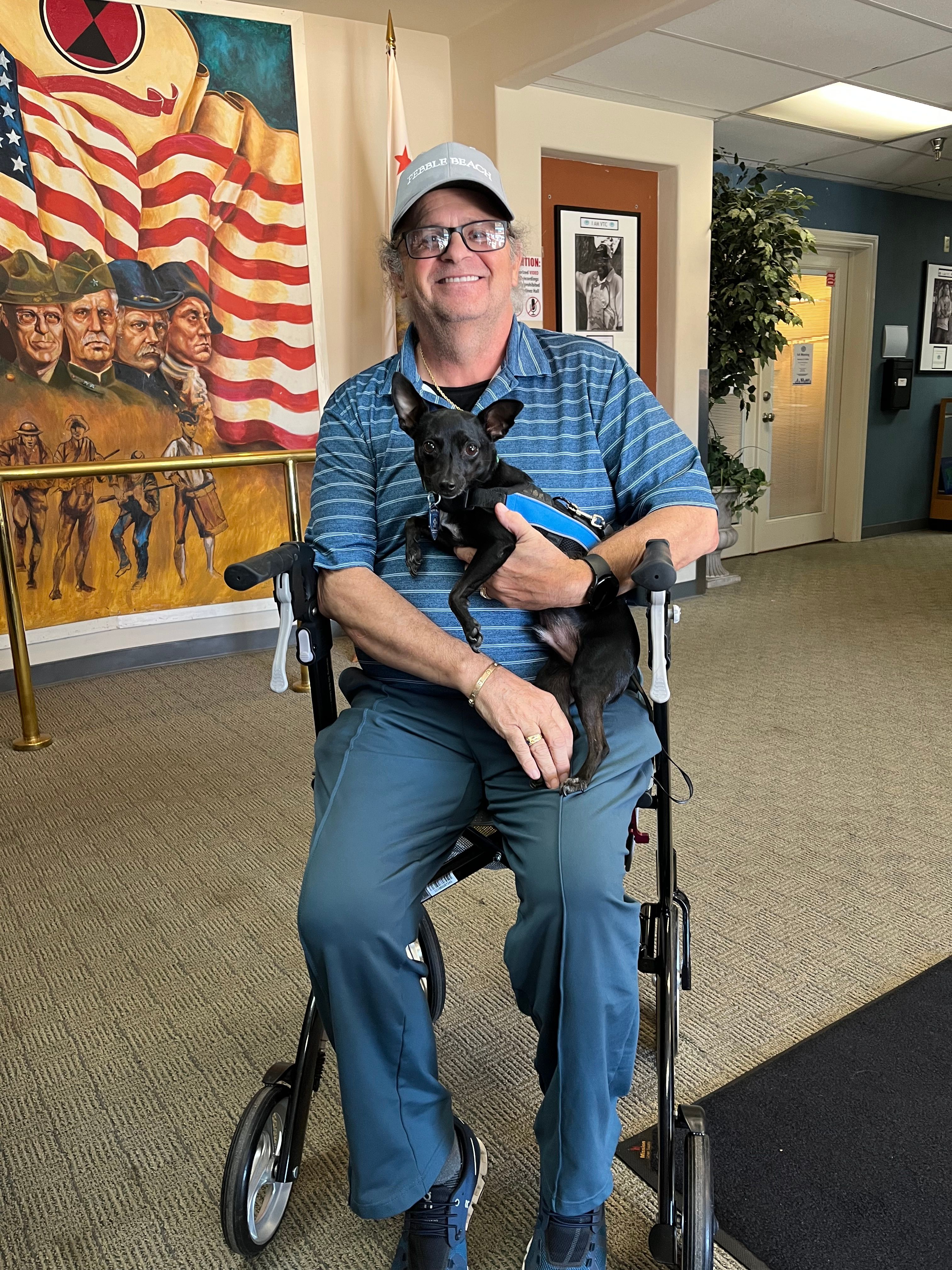 A Veteran seated in a wheelchair holding a small black service dog in the VTC lobby