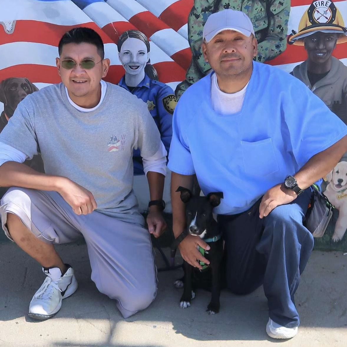 Two Veterans at CTF Soledad sitting in front of a mural, holding a dog from Wiley's Wish