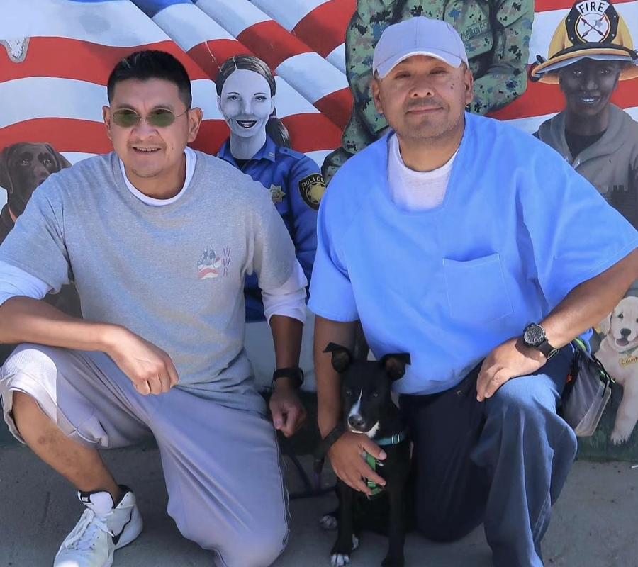 Two Veterans at CTF Soledad sitting in front of a mural, holding a dog from Wiley's Wish