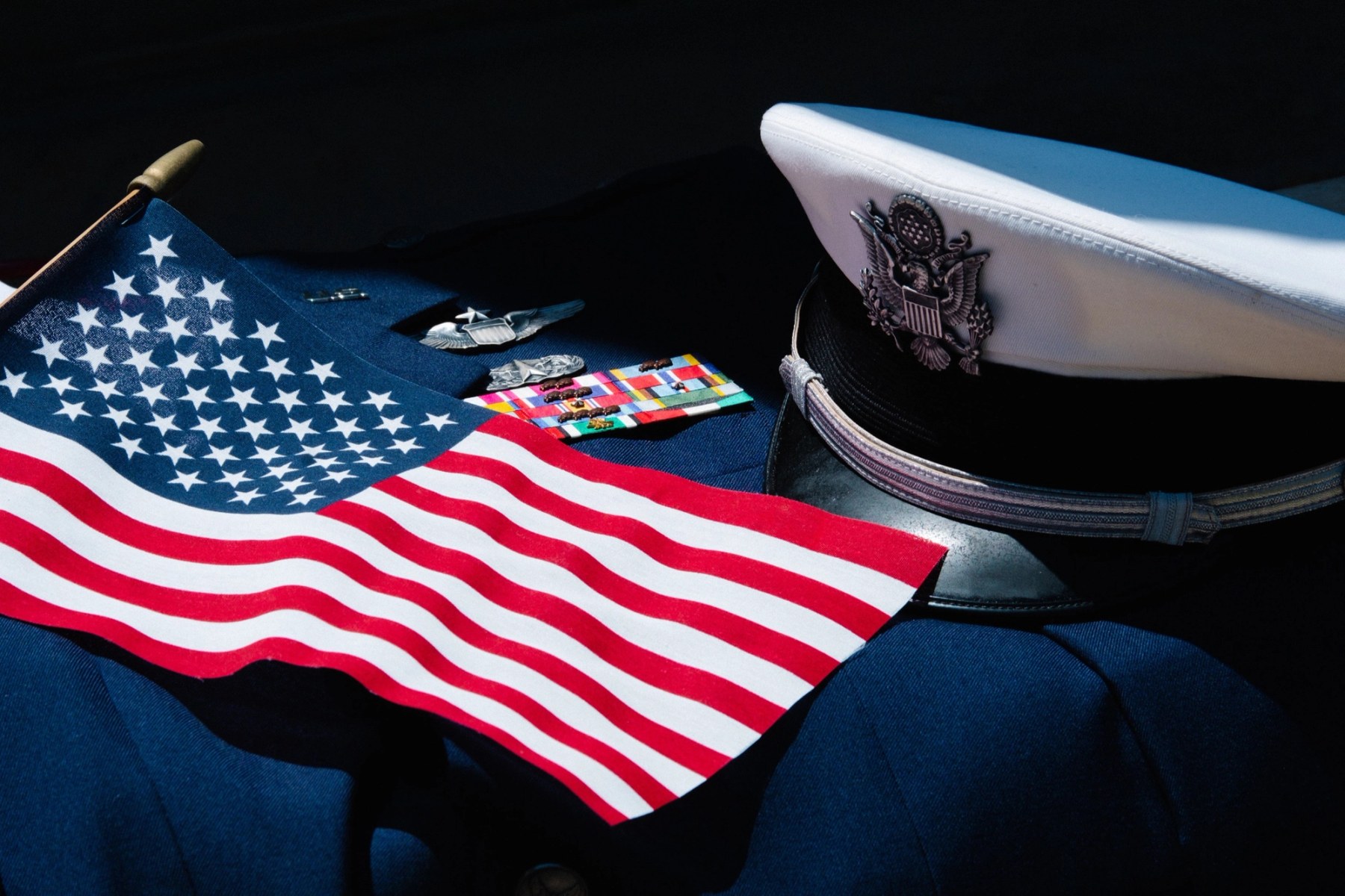 An American flag draped over a folded dress uniform beside a white service cap with ribbons and insignia