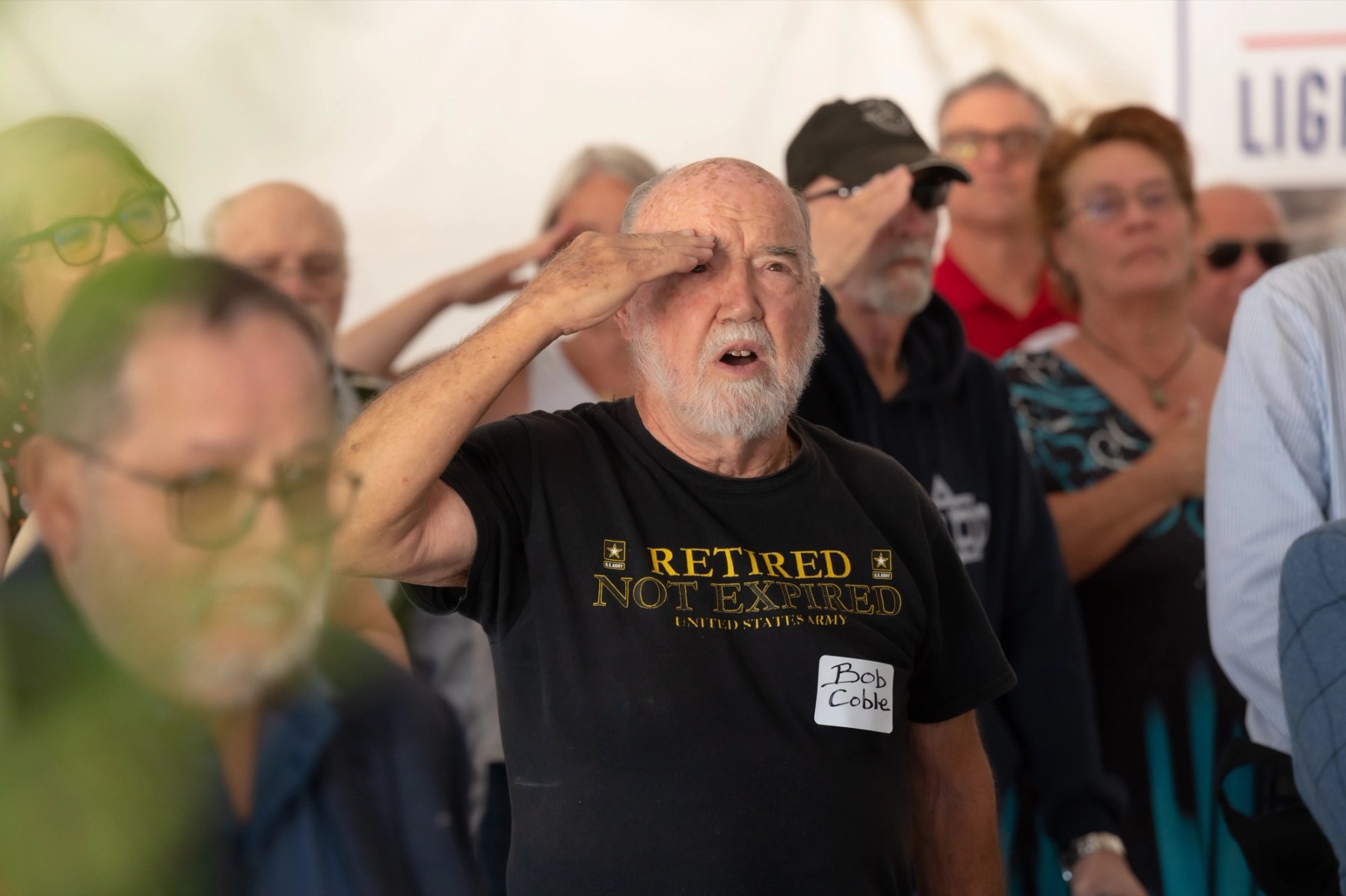 A veteran holds a salute during an outdoor ceremony at Lightfighter Village, with fellow attendees saluting behind him