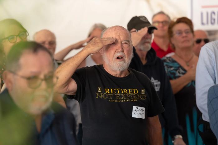 A Veteran holds a salute during an outdoor ceremony at Lightfighter Village, fellow attendees saluting behind him