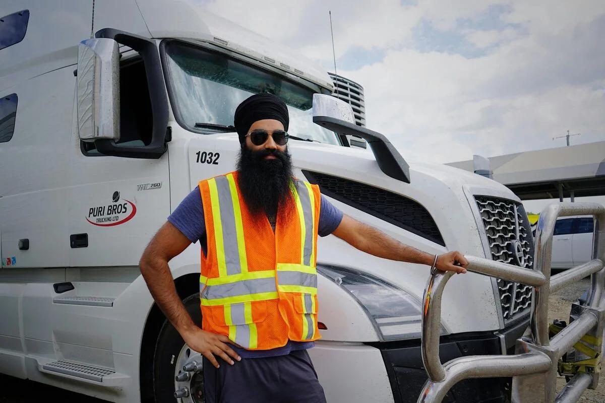 A Sikh commercial truck driver in a safety vest standing beside his semi truck, an authentic messenger in the UNITED SIKHS Freedom Drivers campaign.