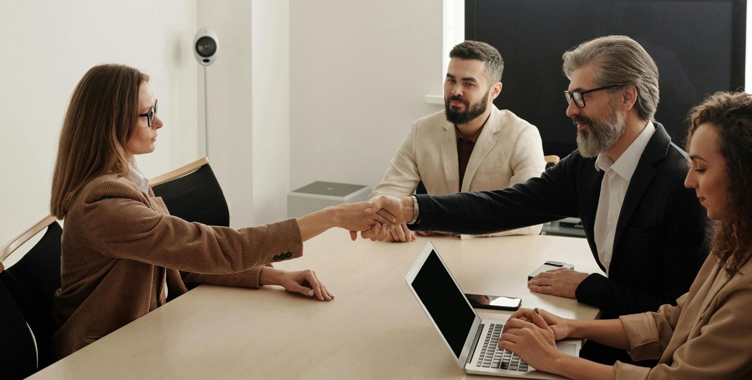 Vier Personen sitzen an einem Tisch, eine Frau und ein Mann geben sich die Hand, eine weitere Frau hat einen Laptop vor sich auf dem Tisch