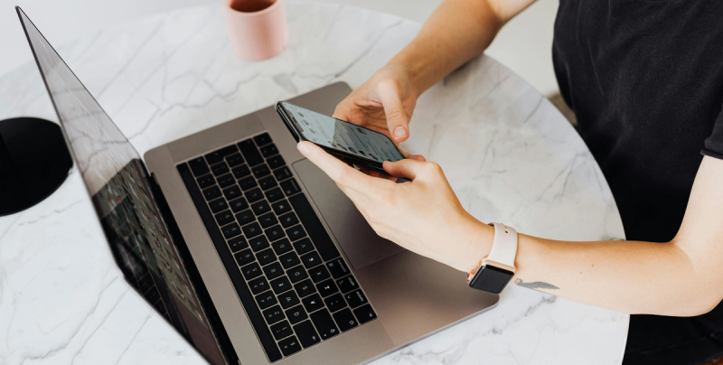 Frau sitzt vor einem Laptop und hält ein Handy in der Hand.