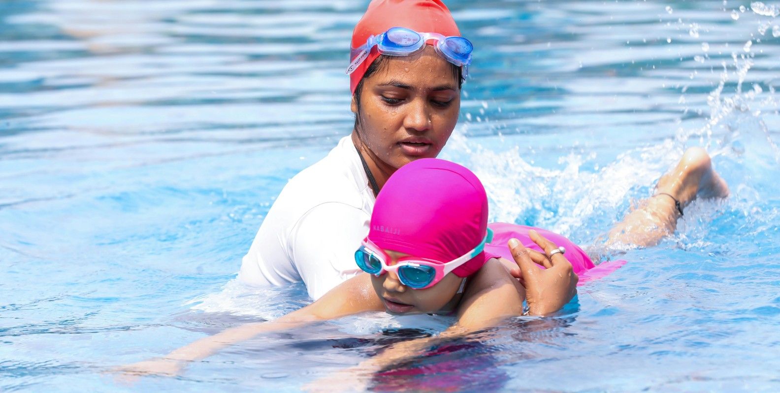 Eine Schwimmtrainerin unterstützt ein Kind beim Schwimmen im Pool. Beide tragen bunte Badekappen und Schwimmbrillen, während das Kind die Schwimmbewegungen übt.