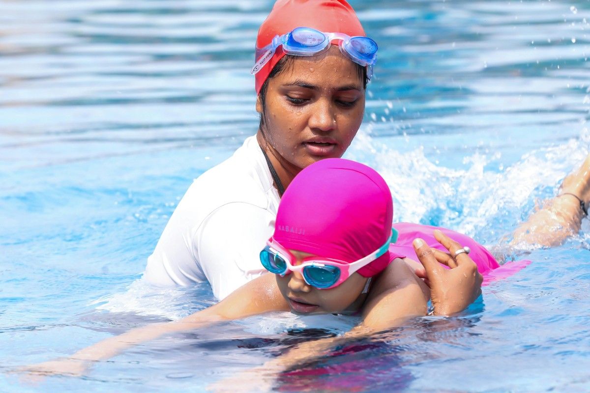 Eine Schwimmtrainerin unterstützt ein Kind beim Schwimmen im Pool. Beide tragen bunte Badekappen und Schwimmbrillen, während das Kind die Schwimmbewegungen übt.