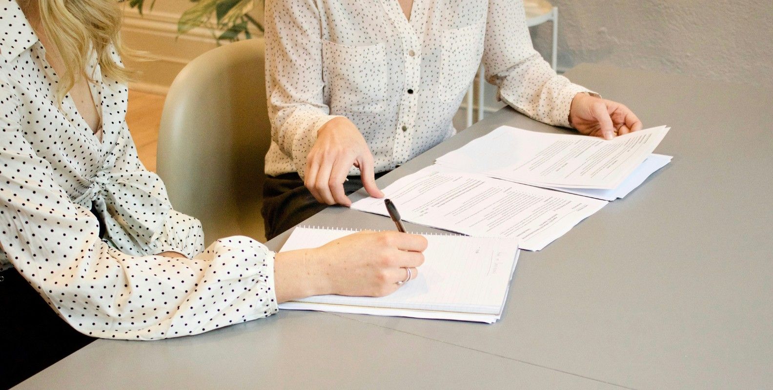 Zwei Personen sitzen an einem Tisch und bearbeiten gemeinsam Unterlagen; eine Person schreibt in ein Notizbuch, die andere zeigt auf Dokumente.