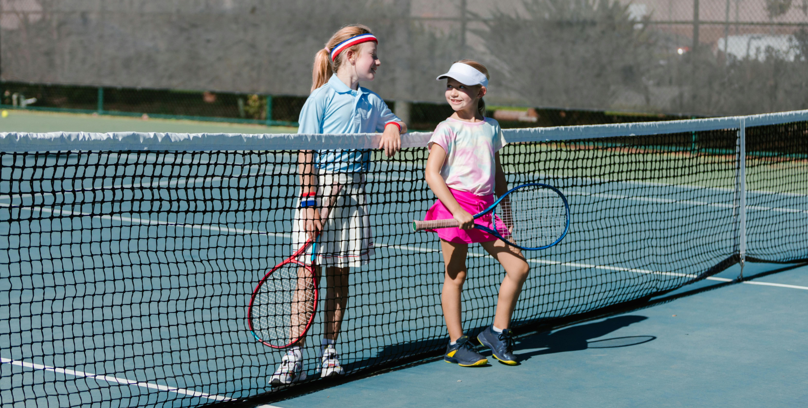Zwei junge Mädchen stehen auf einem Tennisfeld im Verein. Beide halten einen Tennisschläger in der Hand.
