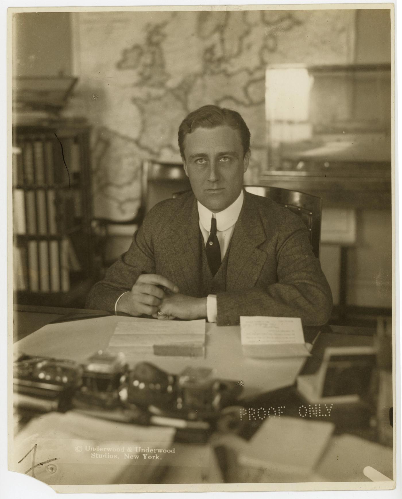 Sepia photograph of a young man in a dark suit seated at a desk with papers and an inkstand, a large map on the wall behind him