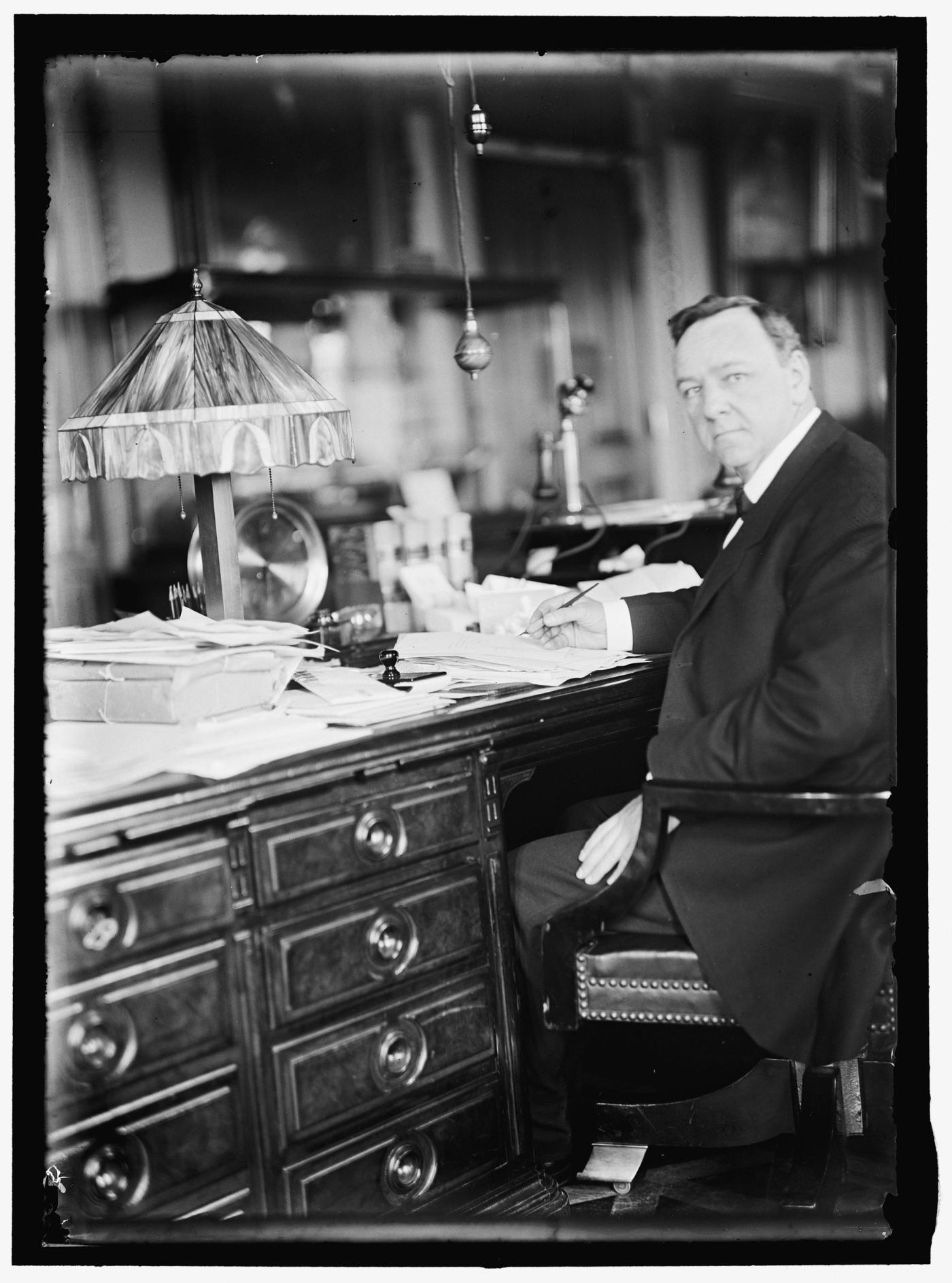 Black-and-white photograph of a heavy-set man in a dark suit seated at a large rolltop desk under a hanging lamp