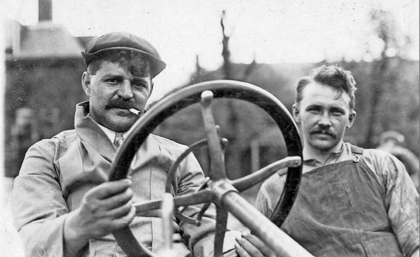 Two men with mustaches posing with an early automobile steering wheel, circa early 1900s.