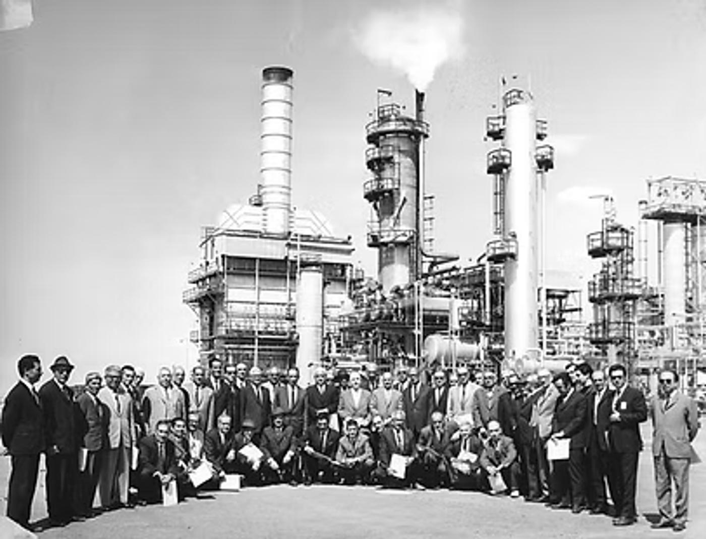 Large group of formally dressed men posed in front of industrial refinery complex with tall smokestacks and processing towers, circa mid-20th century.