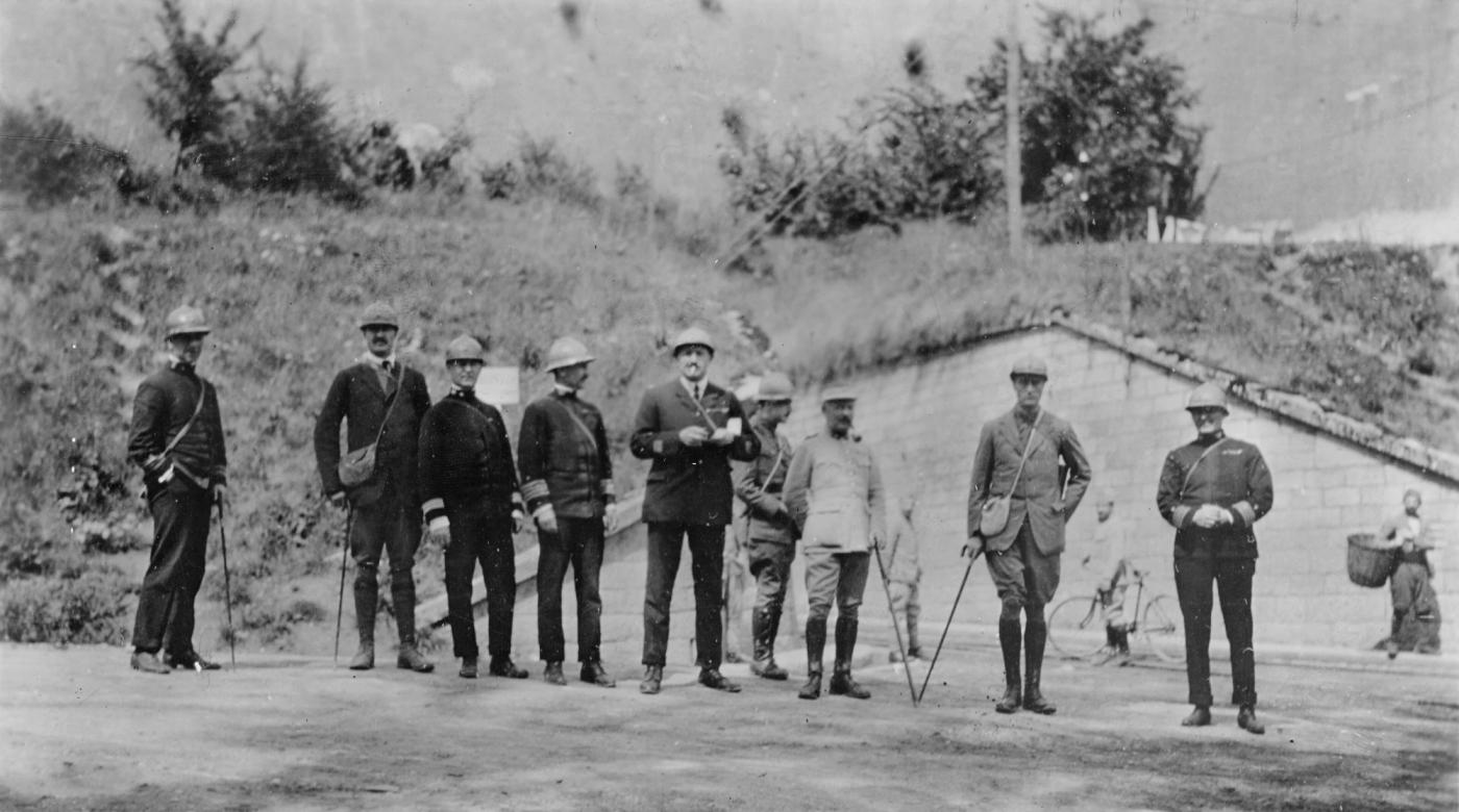 Black-and-white photograph of a group of men in military and civilian clothes, several in steel helmets, standing in a row before a grassy earthwork
