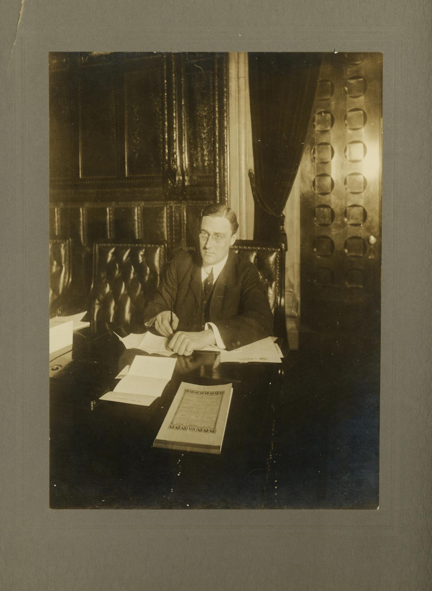 Sepia photograph of a young man in a dark suit seated at a desk with papers in front of a tall paneled wall