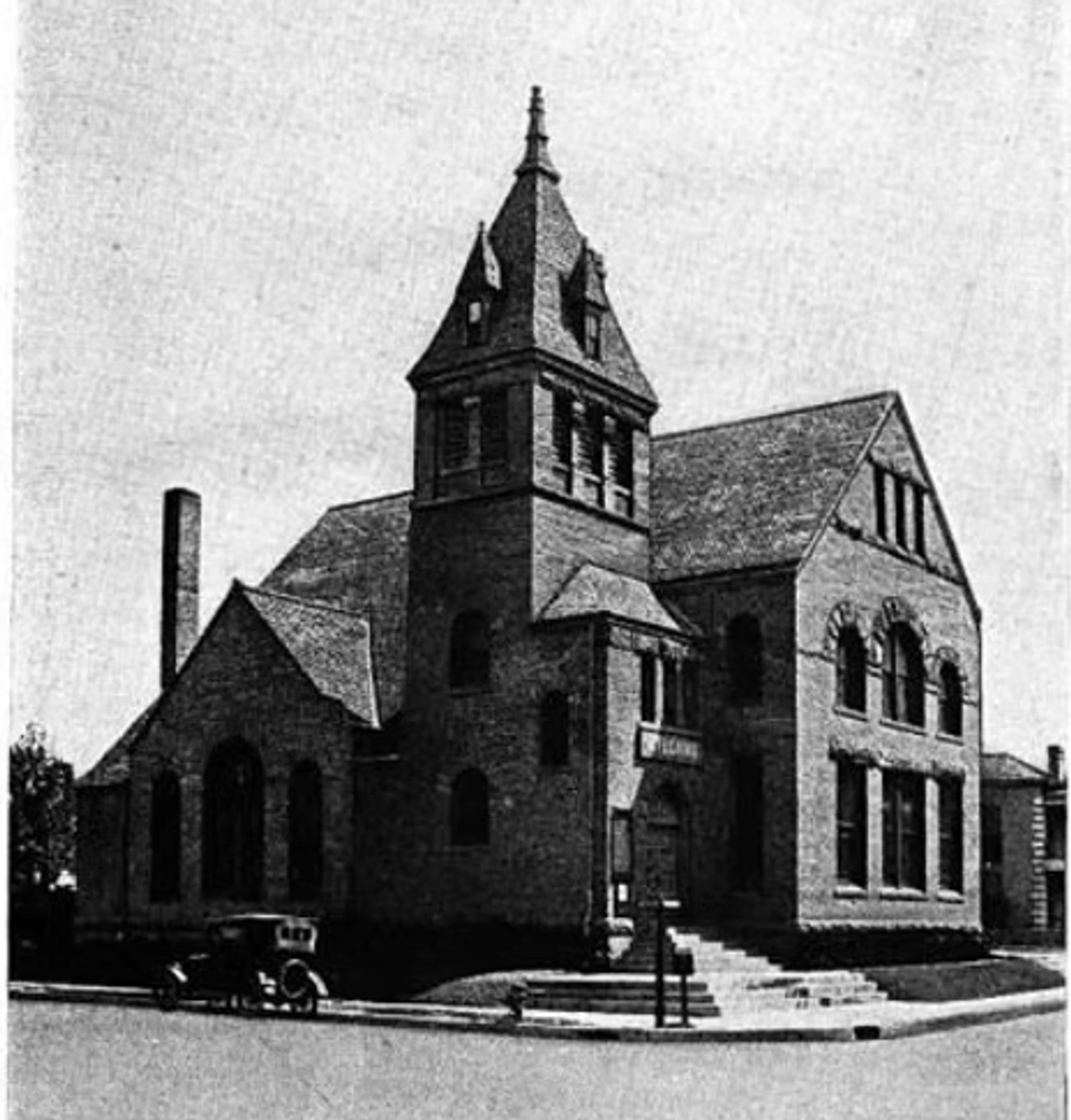 Historic brick church building with tall steeple and vintage automobile parked outside, early 20th century architecture.
