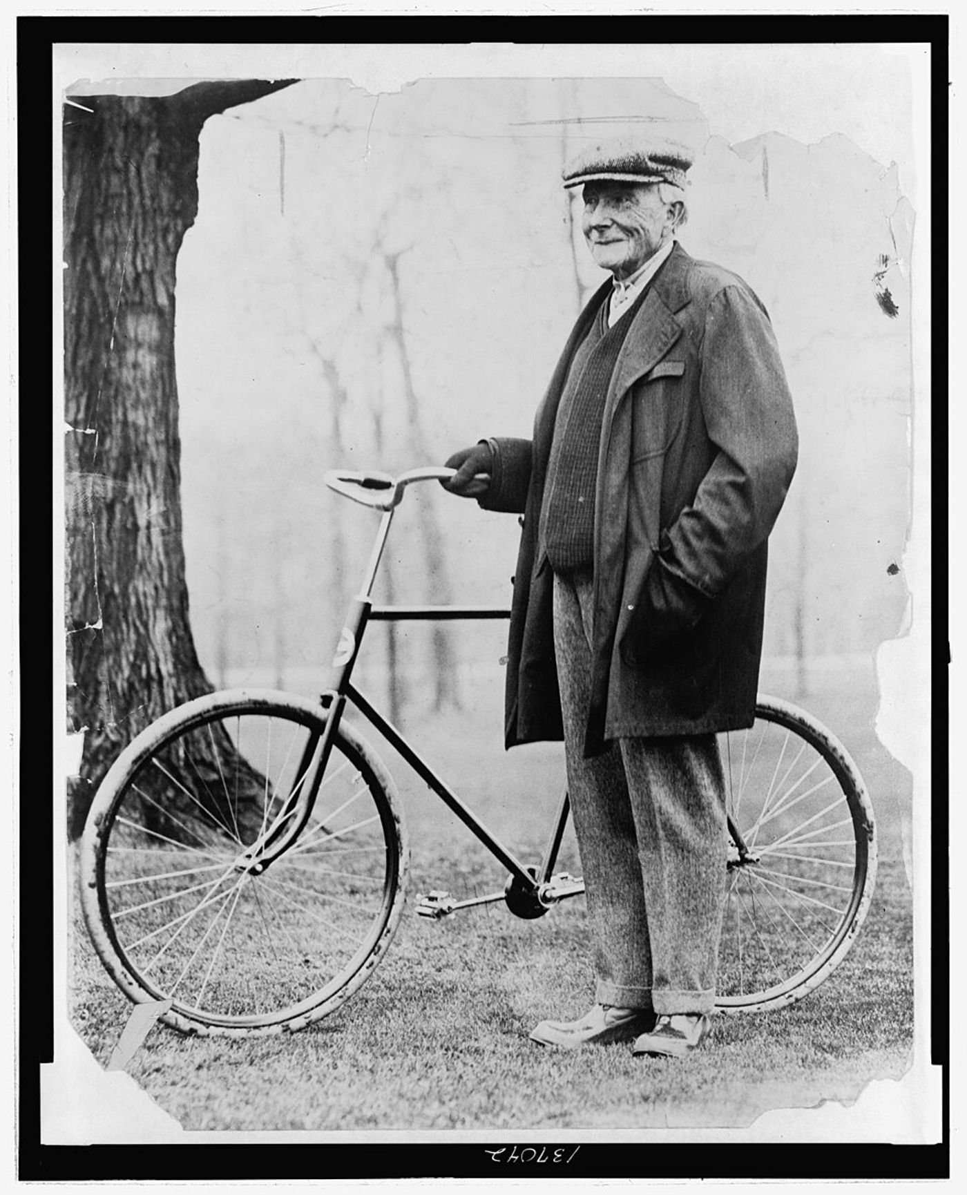 John D. Rockefeller in suit and cap standing with bicycle against white wall and tree trunk.