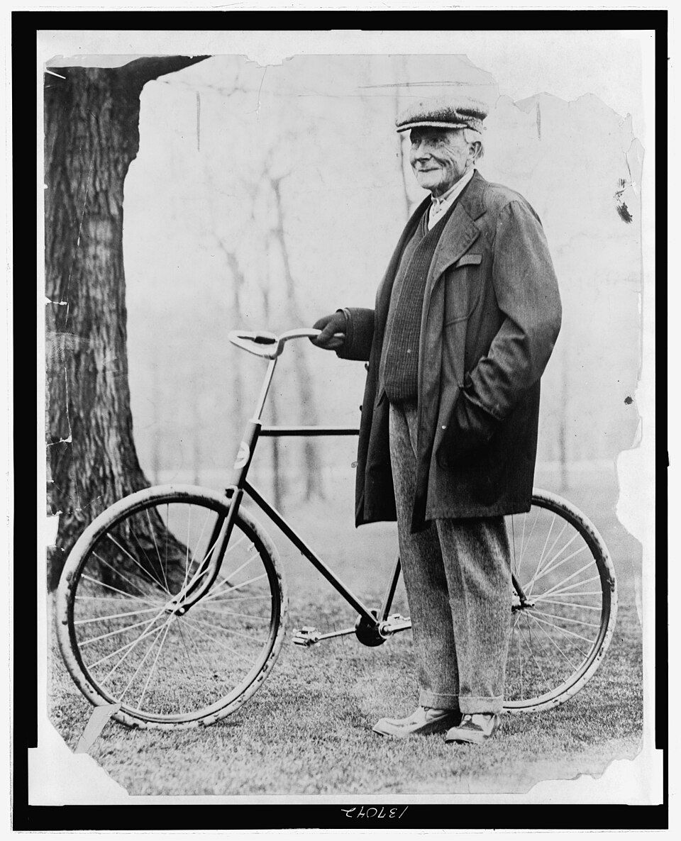 John D. Rockefeller in suit and cap standing with bicycle against white wall and tree trunk.