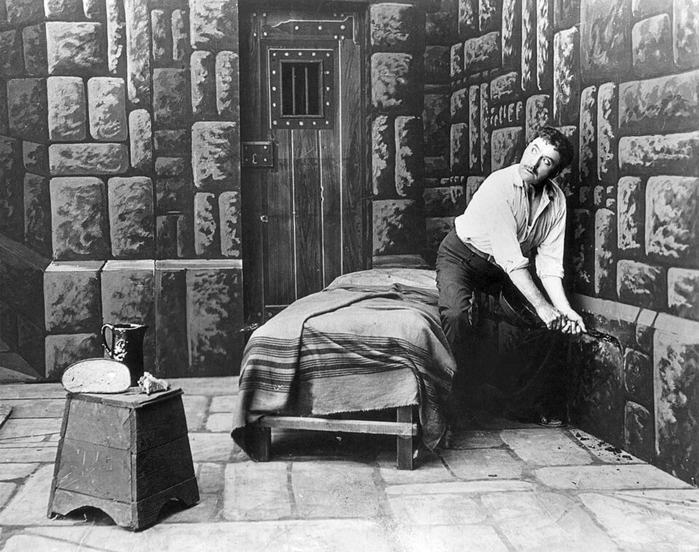 Black and white film scene showing man in white shirt sitting on bed in stone prison cell with barred door.