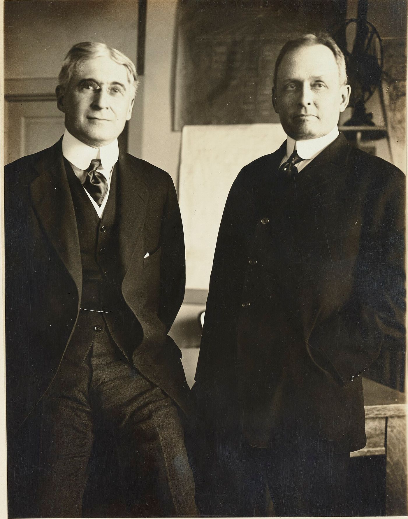 Sepia photograph of two middle-aged men in dark suits standing side by side in an office