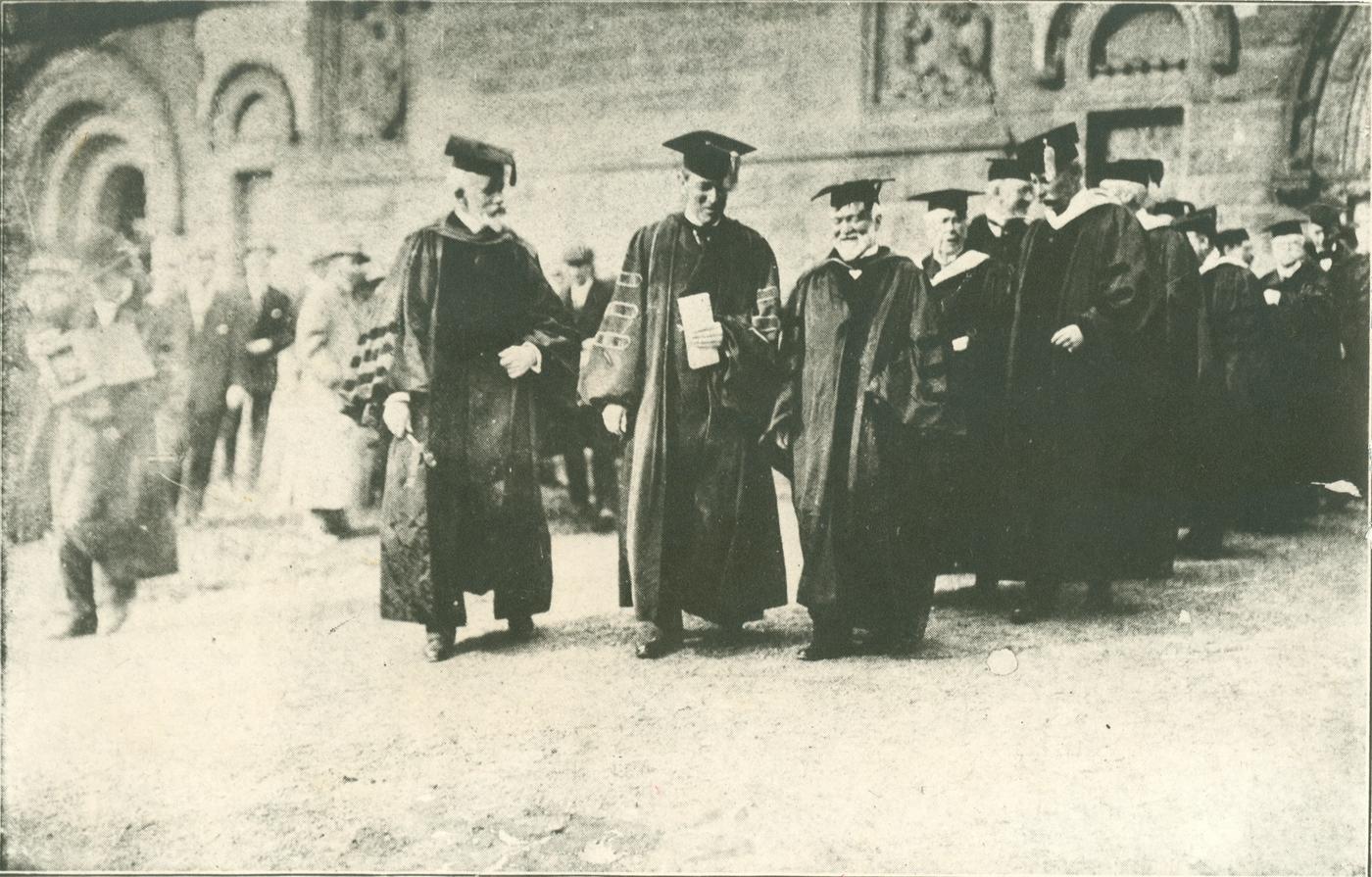 Black-and-white photograph of men in academic caps and gowns walking in procession past a stone building, a younger clean-shaven man at center between two bearded older men