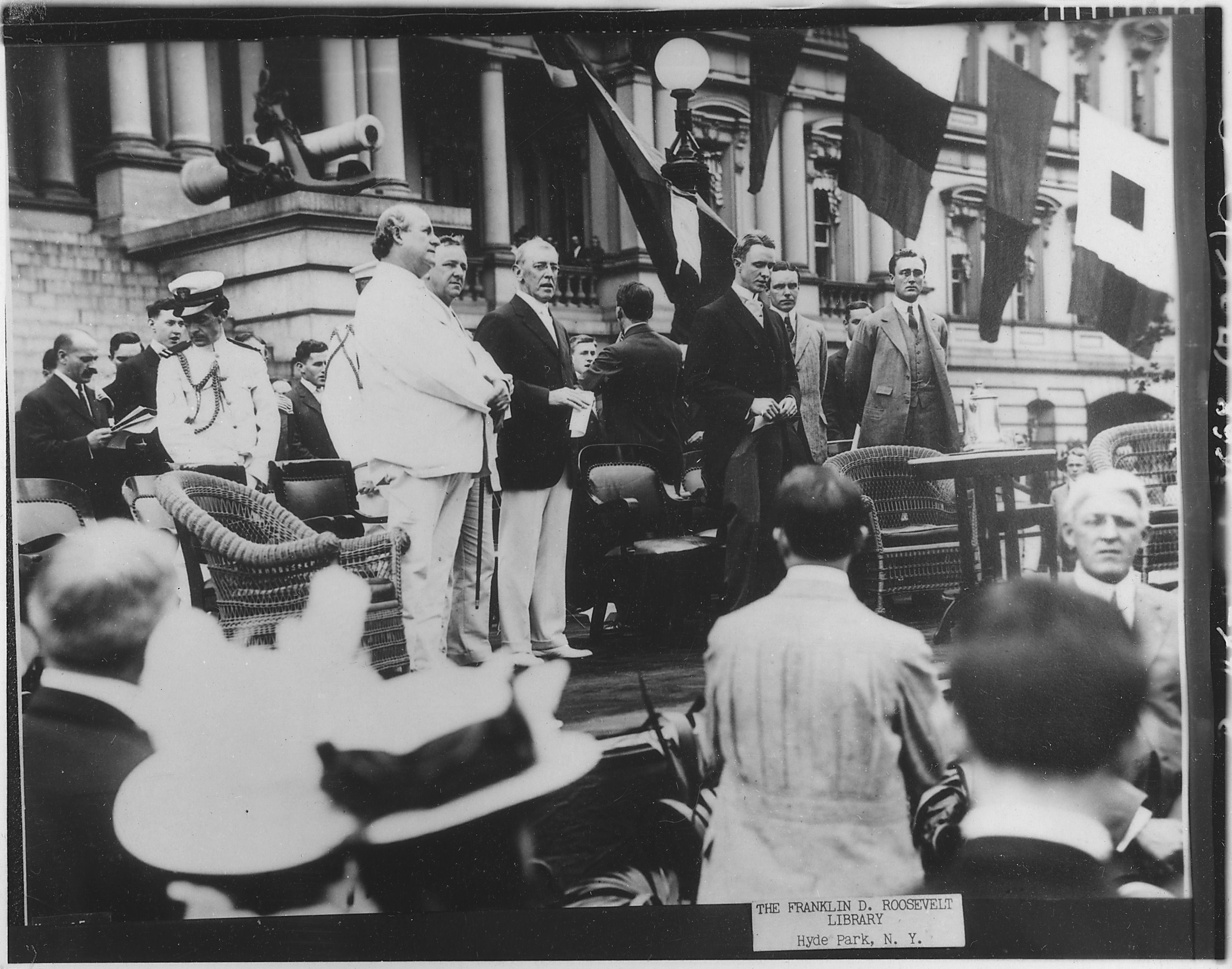 Black-and-white photograph of a crowd gathered at an outdoor Flag Day ceremony in front of a government building hung with flags, with officials standing on a platform