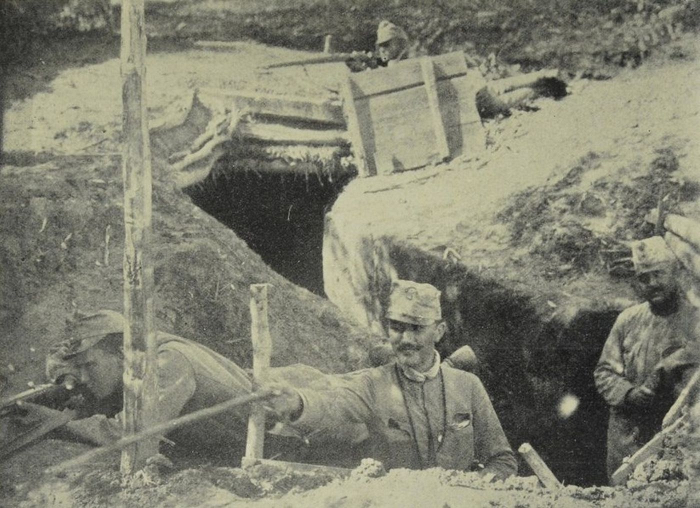 Grainy black-and-white photograph of soldiers in a dugout trench, the man at left in a field cap leaning on the parapet