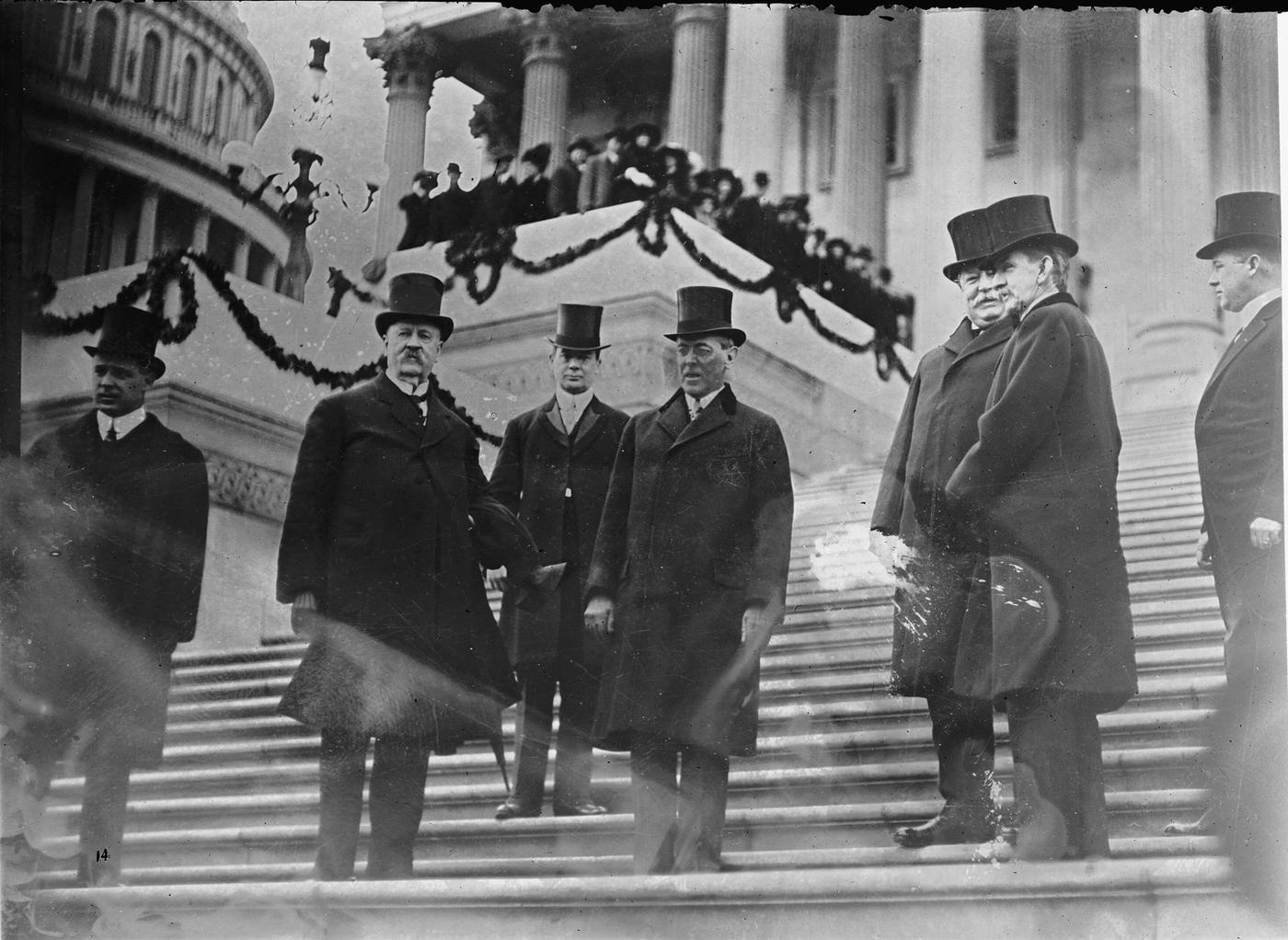 Black-and-white photograph of several men in top hats and long overcoats standing on the broad steps of the Capitol, with a decorated stand and spectators behind them